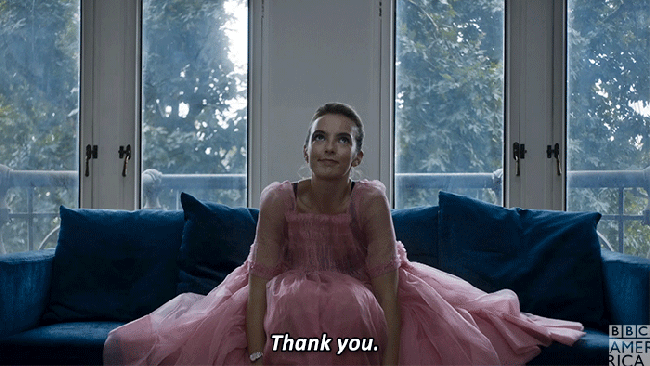 A gif from Killing Eve showing Villanelle sitting on a sofa in a pink dress shrugging and saying thank you.